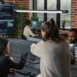 Software programer pointing pencil at source code on computer screen explaining algorithm to coworker standing next to desk. Programmers discussing online cloud computing with team.